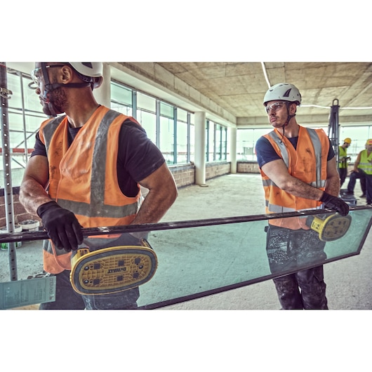 Two construction workers wearing safety helmets and orange vests use DEWALT DCE590 suction tools to carry a large glass