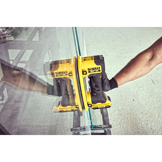 A person wearing gloves is using a yellow DEWALT DCE590 18V XR cordless tool to lift or hold a large glass pane. The tool is