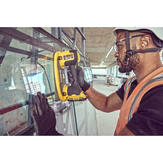 A person wearing gloves and a safety vest uses a DEWALT DCE590 tool to handle a large glass panel at a construction site