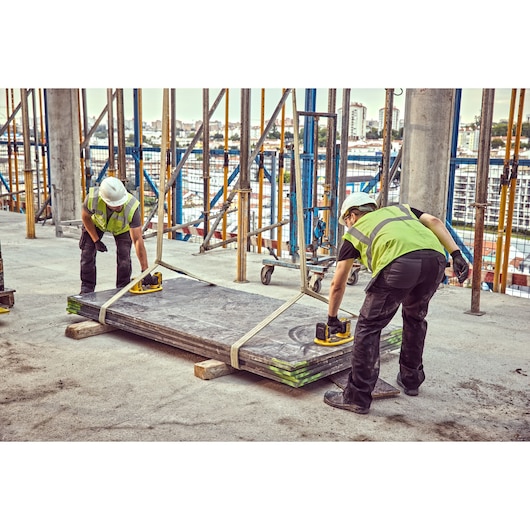 Two construction workers wearing safety vests and helmets use DEWALT cordless tools to lift and position large flat building