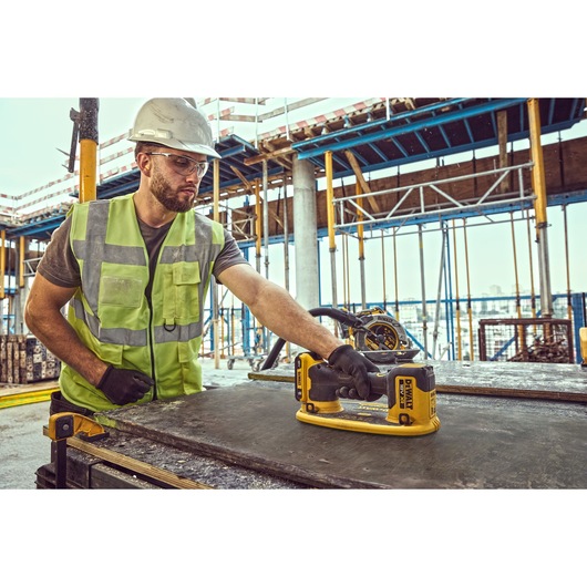 A person wearing a safety helmet and high-visibility vest is using a DEWALT DCE590 power tool on a construction site. The