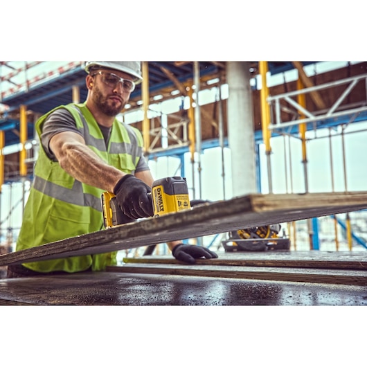 A person wearing a safety vest and gloves using a DEWALT power tool to cut a piece of wood on a construction site, with