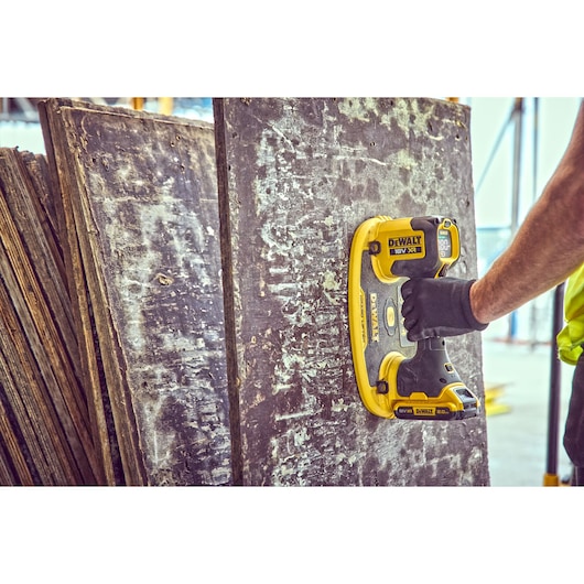 A person wearing a black glove uses a yellow DEWALT 18V XR power tool to sand or clean a large, weathered wooden board at a
