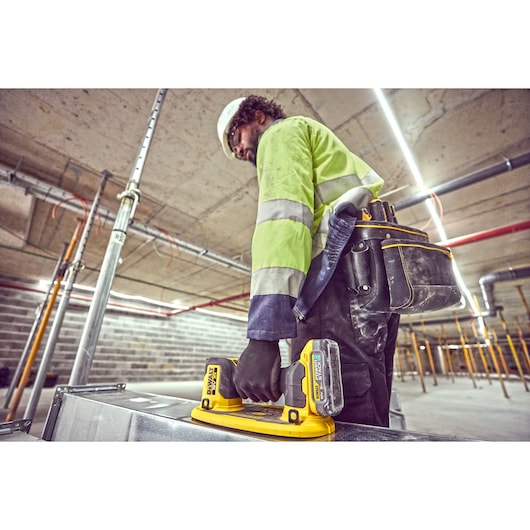 Person wearing a high-visibility jacket and tool belt working in an industrial environment, holding a DEWALT DCE590 tool.