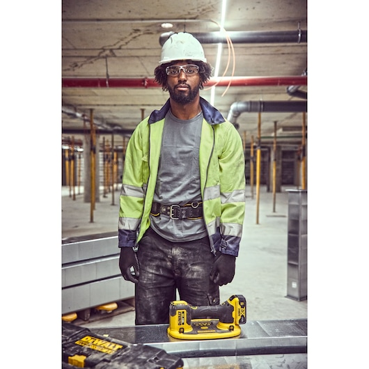 A construction worker wearing a safety helmet, high-visibility jacket, gloves, and a tool belt stands in an indoor