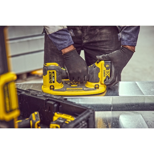 A person wearing black gloves using a DEWALT branded cordless power tool, positioned on a metal surface. Several DEWALT
