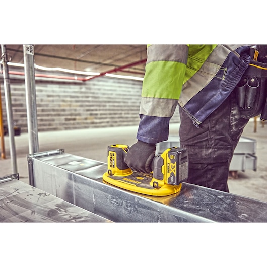 A person wearing a high-visibility jacket and black gloves is using a yellow DEWALT cordless power tool on a metal duct in
