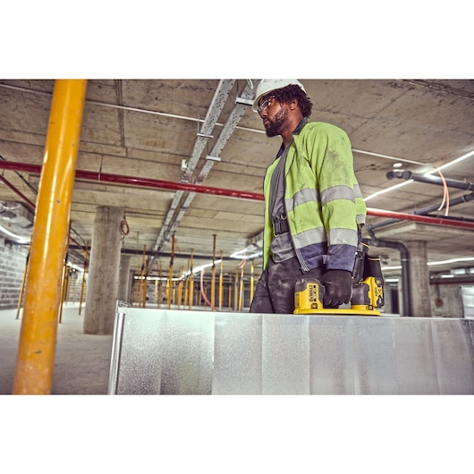 A person wearing a reflective jacket and gloves is standing in a construction site, holding a DEWALT DCE590 cordless tool