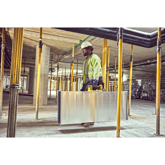 A construction worker wearing a high visibility jacket and hard hat carries a large metal panel using two yellow-handle