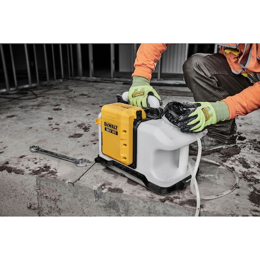 A DEWALT DCE6820N 18V XR industrial photography-related device is being operated by a person wearing green gloves and an orange shirt. The device is placed on a concrete surface at a construction site.