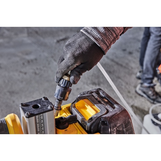 A close-up of a gloved hand adjusting a component on a DEWALT tool, with a yellow and black casing visible. The scene appears to be set in a workshop or construction site.