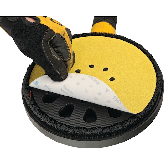 A close-up view of a gloved hand attaching or removing a circular sanding disc from a power tool, with the disc featuring yellow abrasive material and multiple holes.