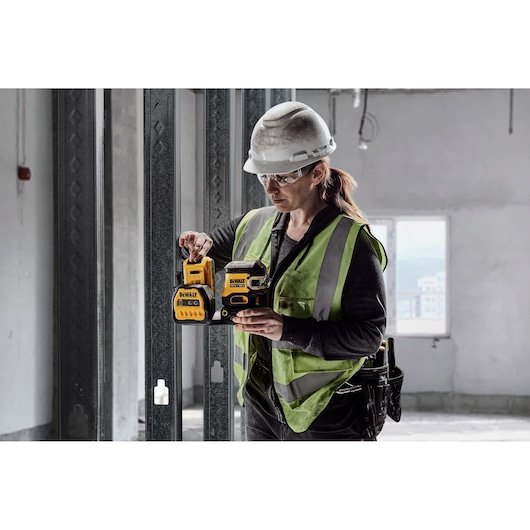 A person wearing a safety vest and hard hat uses a DEWALT green laser level (model DCE822D1G18) on a construction site. The background shows an unfinished building interior with metal wall frames.