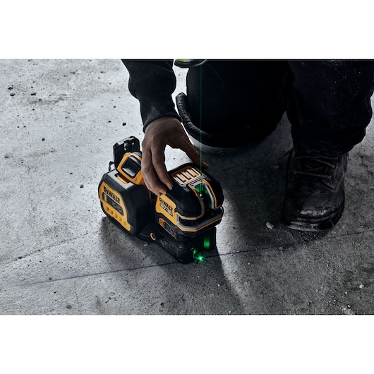 A person is operating a DEWALT laser level tool on a concrete floor, aligning it with a green laser line for precise