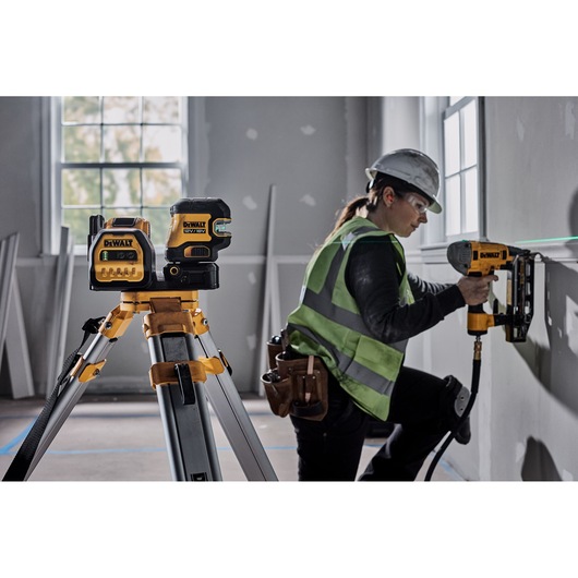 A DEWALT laser level device is mounted on a tripod in the foreground, while a construction worker wearing a safety vest and