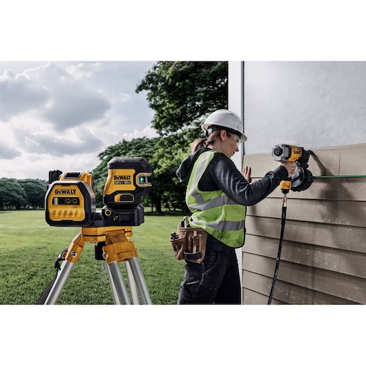 A DEWALT laser level mounted on a tripod is being used outdoors by a construction worker wearing a safety helmet and