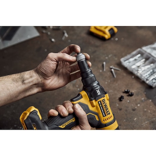 A person is holding and adjusting a DEWALT cordless power tool with a yellow and black design on a workbench, surrounded by screws and small parts.