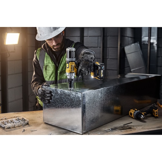 A person wearing protective gear uses a DEWALT DCF414NT cordless power tool to work on a large metal box in an industrial setting. Various tools and screws are visible on the worktable.