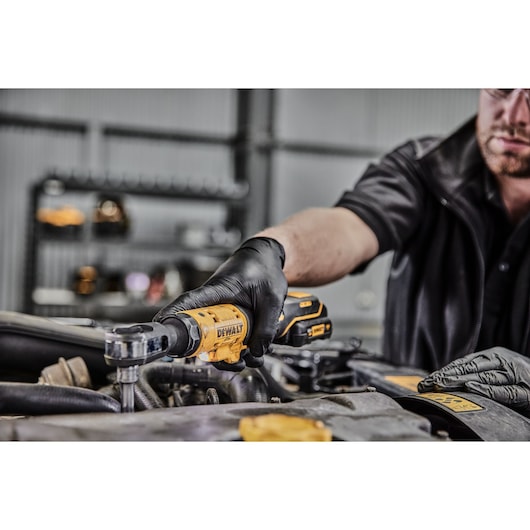 A person wearing black gloves uses a yellow DEWALT cordless ratchet tool to work on a car engine in a workshop setting.
