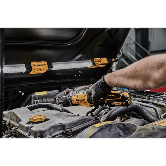 A person using a DEWALT cordless ratchet tool to work on a car engine, with a DEWALT work light attached to the underside of the car hood.