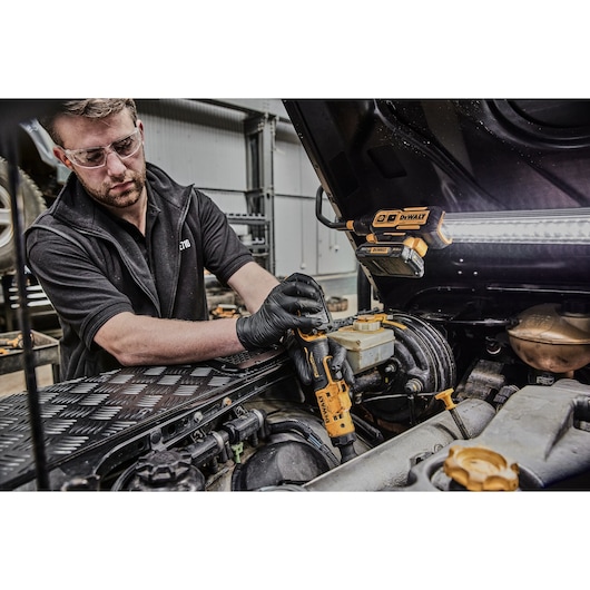 A person wearing black gloves uses a DEWALT DCF504L1G cordless tool to work on a vehicle engine. A DEWALT work light is mounted under the open hood. The scene is in an automotive workshop.