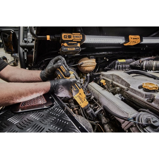 A person wearing black gloves uses a DEWALT cordless ratchet tool to work on a car engine. Additional DEWALT lighting equipment is attached to the open hood for better visibility.