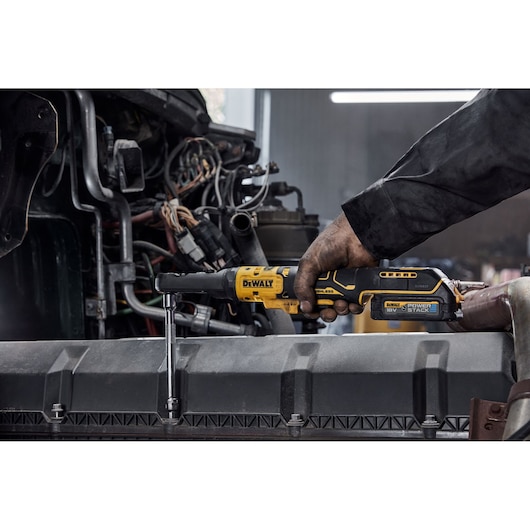 A person using a DEWALT 18V cordless ratchet wrench on an engine part in a workshop. The DEWALT tool features a Powerstack