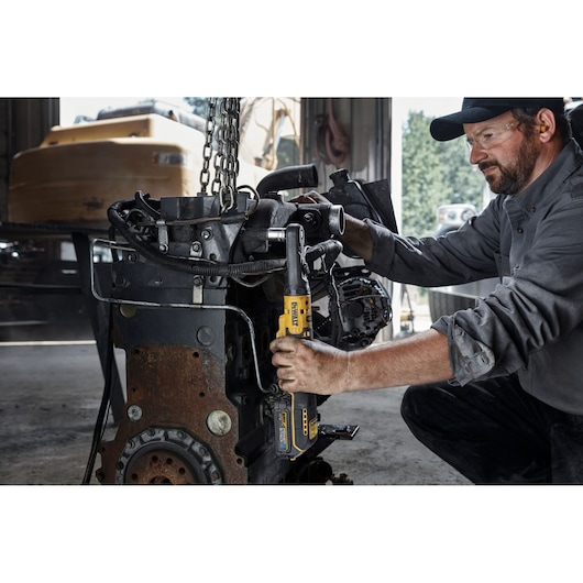 A person using a DEWALT cordless ratchet tool (model DCF510E2G) to work on a large industrial engine in a workshop. The