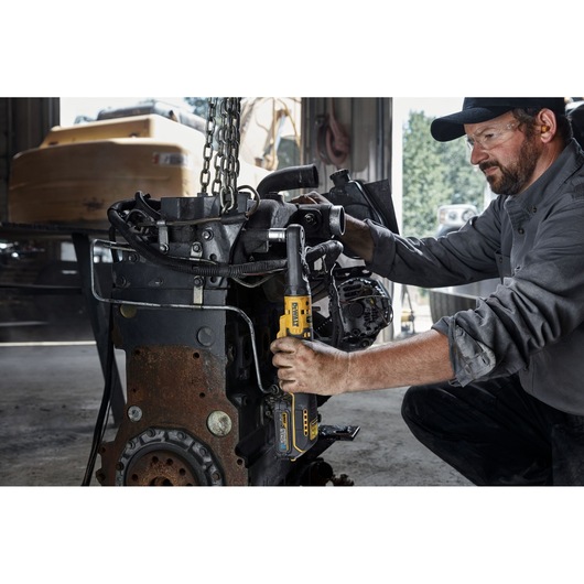 A person using a DEWALT DCF510N cordless ratchet tool to work on an engine inside a workshop. The person's face is