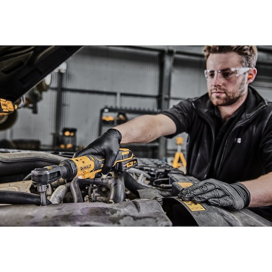 A person wearing black gloves uses a DEWALT DCF512N cordless ratchet tool to work on the engine of a vehicle. Their face is