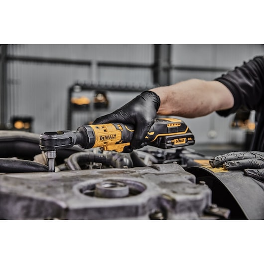A person wearing black gloves uses a DEWALT cordless ratchet wrench to work on an engine part in a workshop.