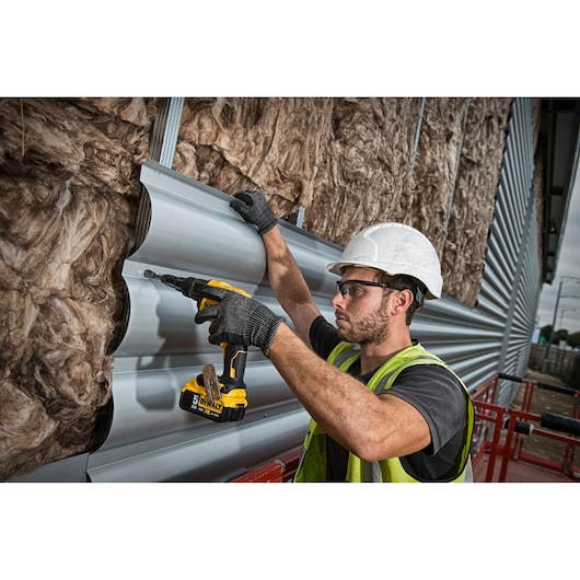 A construction worker wearing a white hard hat and gloves uses a DEWALT DCF622N cordless screwdriver to fasten metal panels onto an insulated wall at an industrial site.