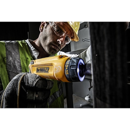 A person wearing gloves and a safety vest uses a yellow DEWALT electric screwdriver (model DCF680G2) with an illuminated tip, working on a piece of equipment in an industrial setting.