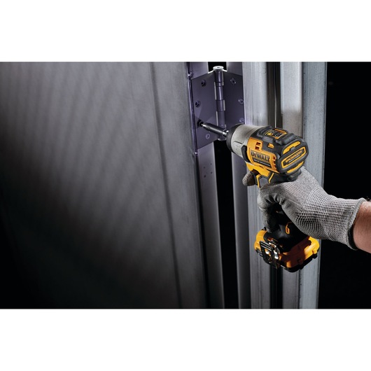 A person wearing a grey work glove uses a DEWALT cordless impact driver to install or tighten screws on a metal door hinge.