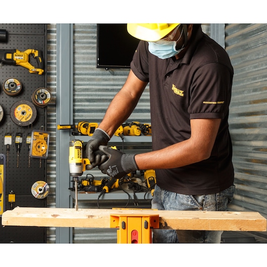 A person wearing safety gear, including gloves, a mask, and a yellow hard hat, is using a DEWALT cordless drill to work on a piece of wood. The background shows various DEWALT tools and accessories organized on a wall and shelves in a workshop setting.