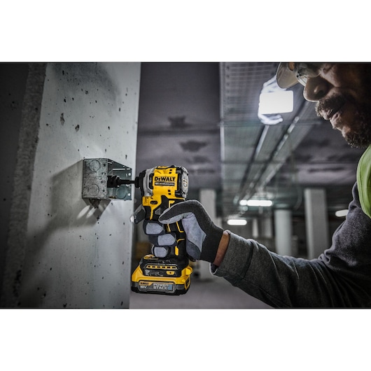 A person wearing gloves uses a DEWALT 18V cordless impact driver to fasten a metal bracket onto a concrete surface in an industrial setting.