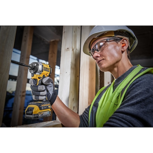 A person wearing a safety helmet and high-visibility vest is using a DEWALT DCF850E1T cordless impact driver to fasten a screw into wooden beams at a construction site. Their face is blurred for privacy.