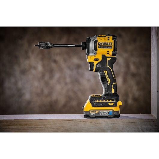 A DEWALT cordless 18V brushless impact driver with a drill bit attached and a POWERSTACK battery, standing upright on a wooden surface.