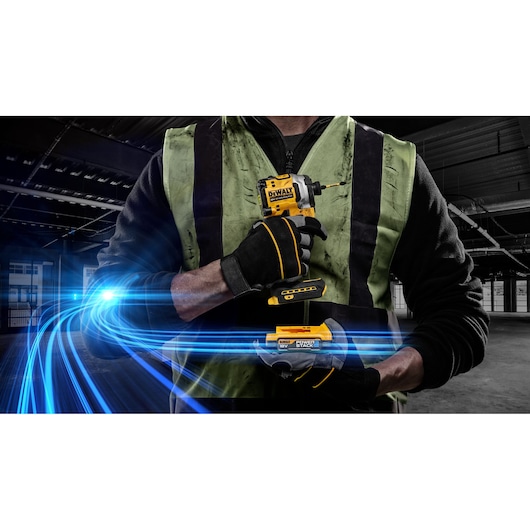 A construction worker wearing a green safety vest and black gloves is holding a yellow DEWALT cordless impact driver with a battery in a dimly lit industrial setting. Blue light streaks are shown in the foreground for a dynamic effect.