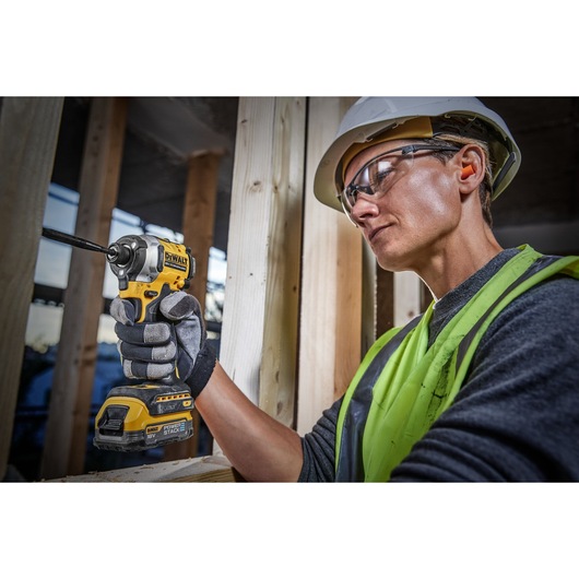 A person wearing a safety helmet, neon safety vest, and gloves uses a DEWALT DCF850E2T 18V cordless impact driver to work on