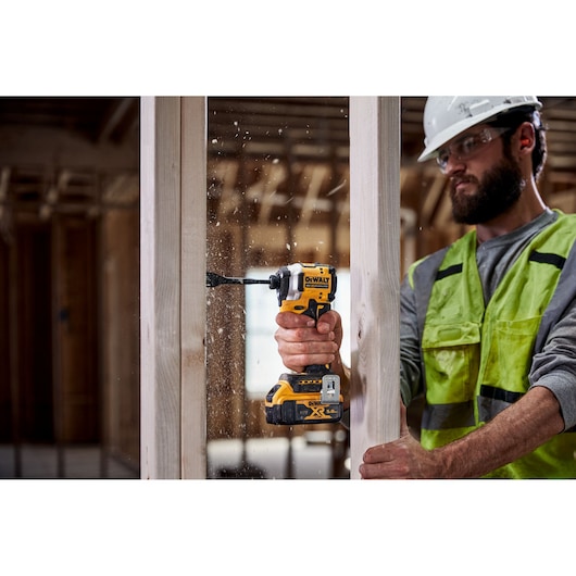 A person wearing a high-visibility vest and a hard hat is using a DEWALT cordless impact driver to drill into a wooden beam