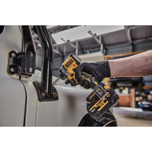 A person wearing a black glove is using a DEWALT DCF850NT cordless impact driver to fasten screws on a vehicle door hinge.