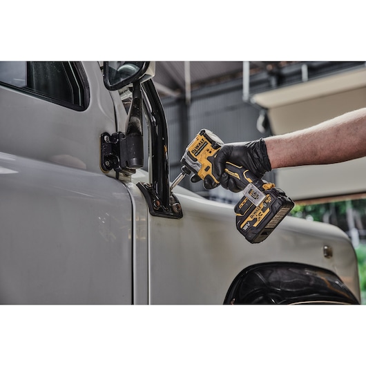 A person wearing a black glove uses a DEWALT DCF850NT cordless impact driver to work on the hinge area of a vehicle door.