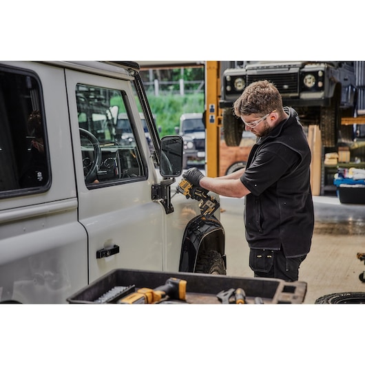 A person is using a DEWALT DCF850N power tool to work on the side mirror of a white vehicle in a garage or workshop. Various other tools are visible in the foreground.