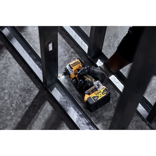 A gloved hand using a DEWALT DCF850P2T cordless impact driver to drill into a metal frame, with the tool's battery visible.