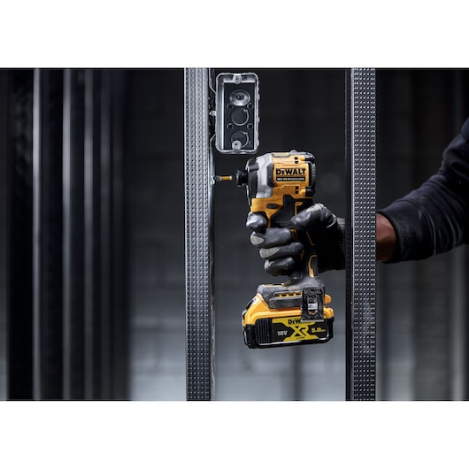 A person wearing gloves uses a DEWALT DCF850P2T 18V XR brushless impact driver to fasten a screw into a metal frame.