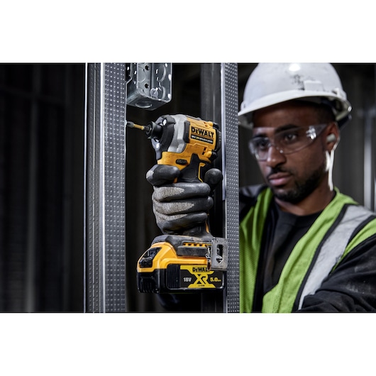 A person wearing safety gear is using a DEWALT DCF850P2T 18V cordless impact driver to fasten screws on a metal frame in an