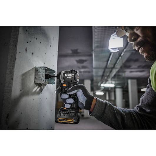 A person wearing gloves and safety gear uses a DEWALT power tool to fasten a metal bracket to a concrete wall inside an industrial building.