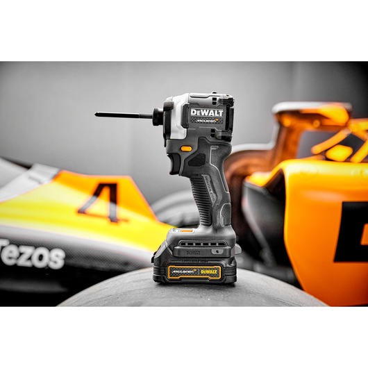 A DEWALT cordless impact driver tool is shown standing upright on a tire, with a racing car featuring orange and yellow details in the background.