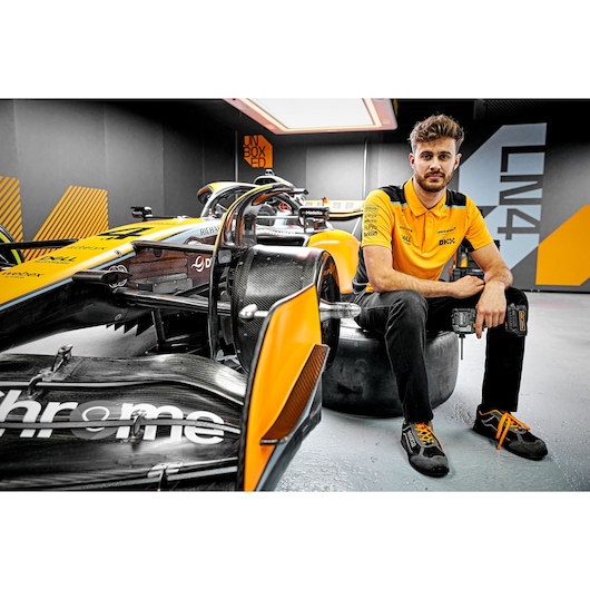 A person in a yellow and black shirt sitting on a tire next to a yellow and black Formula 1 race car inside a modern garage. The person is holding a DEWALT power tool. The person's face is blurred.